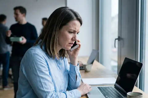 Symbolbild: Eine Frau telefoniert besorgt am Fenster ihres Büros, KI-generiert