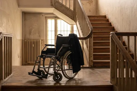 Symbolbild: Ein leerer Rollstuhl steht im Treppenhaus eines Altbaus vor der Treppe nach oben, KI-generiert