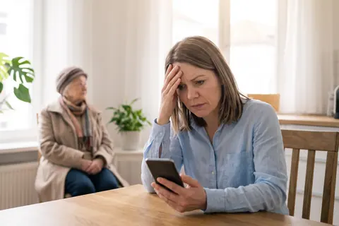 Symbolbild: Eine Frau hält sich besorgt die Stirn und schaut auf ihr Telefon, im Hintergrund wartet eine ältere Frau im Mantel, KI-generiert